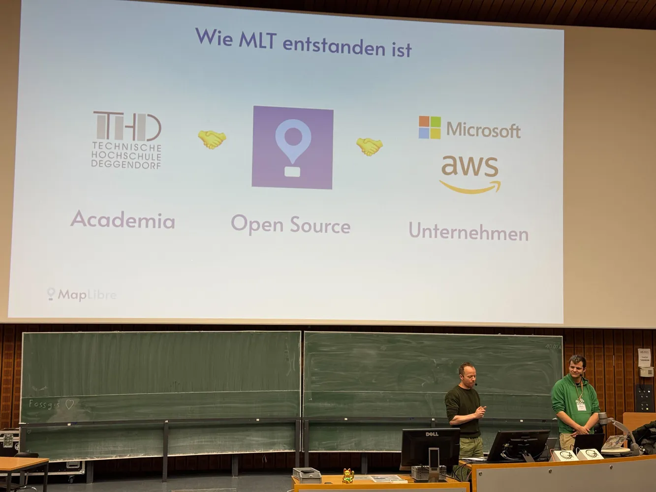 Photo from Frank's and Bart's talk on MapLibre at FOSSGIS. On the slide: Academia, Open Source and Organizations, with hand shaking emoji between them.
